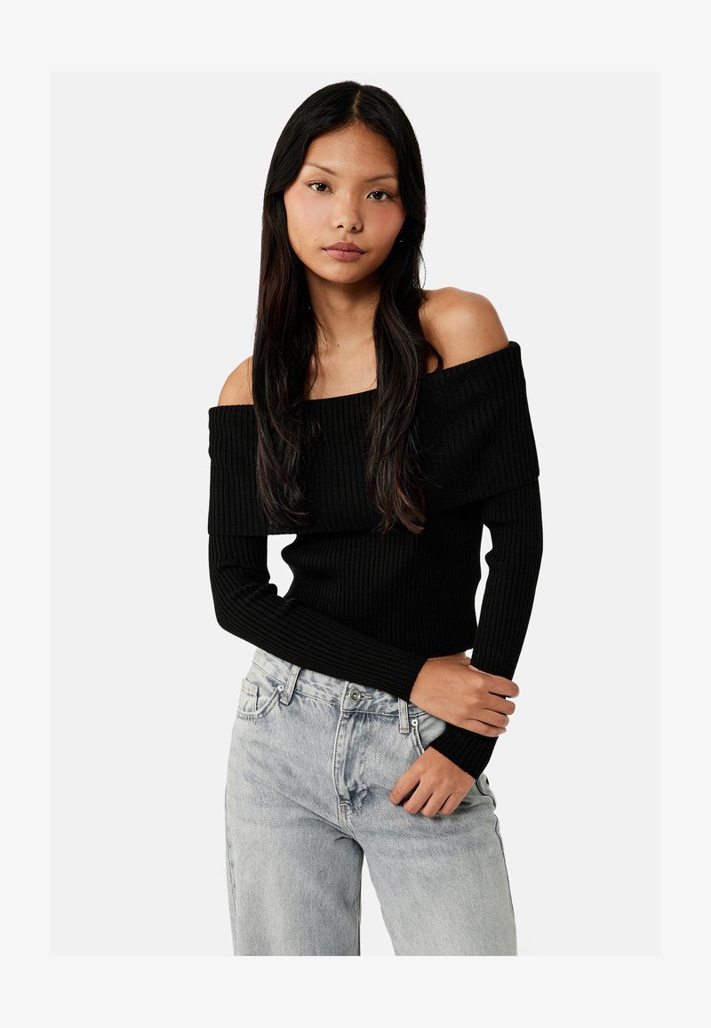 Ung kvinde med langt mørkt hår iført en sort off-shoulder ribbet sweater og lysegrå high-waist jeans, poserer mod en ensfarvet baggrund.