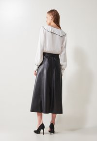 Gonna midi in pelle nera con una texture liscia e cuciture sottili, abbinata a una blusa bianca con colletto e polsini arricciati a contrasto.
