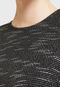 Czarny top wykonany z teksturowanej, metalicznej dzianiny z nieregularnymi białymi paskami. Okrągły dekolt i bezrękawnik, podkreślający połysk tkaniny.