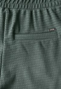 Groene textuur broek met een geribde tailleband, voorzien van een achterzak en een klein zilveren logolabel. De stof heeft een geruit patroon.