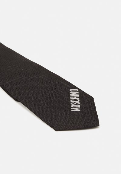 Svart fluga med en texturerad yta, med ordet "MOSCHINO" broderat i vitt på ena sidan. Månghörnig spet design.