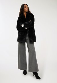 Czarny płaszcz z sztucznego futra z kołnierzem typu szal, charakteryzujący się miękką teksturą i luźnym krojem, zestawiony z szerokimi szarymi spodniami i czarnymi szpilkami w szpic.