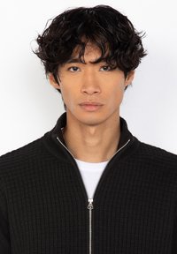 Czarna dziana kurtka o fakturze z kołnierzem stojącym i zamkiem błyskawicznym z przodu. Nosi ją osoba w białej koszuli, a zdjęcie ukazuje zbliżenie na twarz modela.