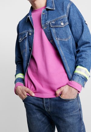 Mann trägt blaue Jeansjacke und Jeans mit neongrünen Streifendetails an den Ärmeln und ein knallpinkfarbenes Langarmshirt, Hände in den Hosentaschen.