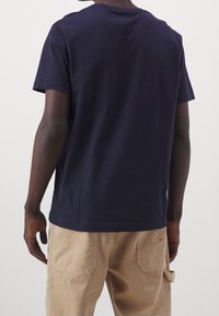 Camiseta de algodón en navy oscuro con mangas cortas y cuello redondo. La vista trasera muestra líneas limpias y un corte relajado, combinada con pantalones beige.