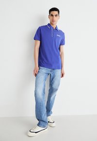 Modrá polo tričko s golierom, s kontrastnými bielymi pruhmi a detailom loga. V parku so svetlomodrými džínsy a bielymi teniskami.