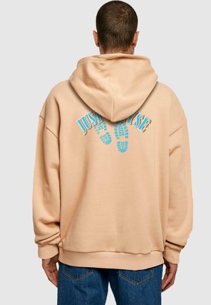 Beige oversized hettegenser med stor blå grafisk tekst "JUST" og fotavtrykkdesign på baksiden. Myk tekstur og justerbar hette.