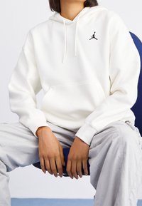 Sudadera blanca con bolsillo tipo canguro. Presenta un logo Jumpman negro en el pecho. Se lleva sobre pantalones gris claro. Tejido suave y corte relajado.
