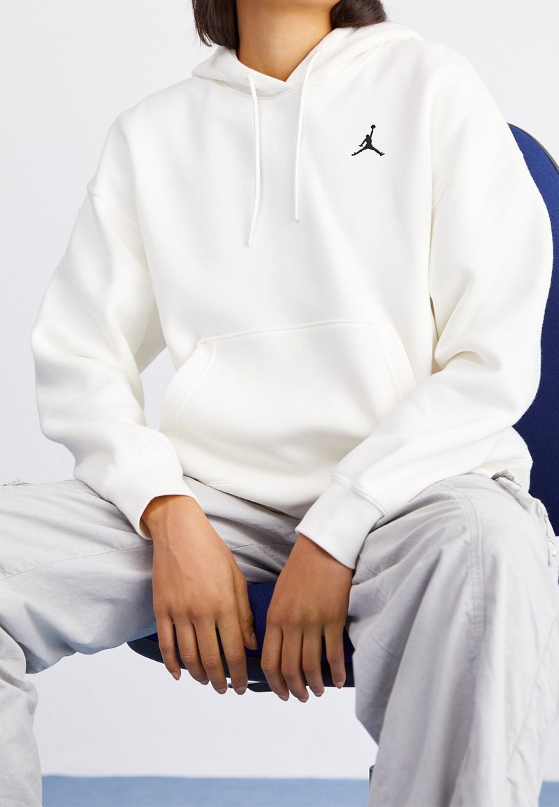 Sudadera blanca con bolsillo tipo canguro. Presenta un logo Jumpman negro en el pecho. Se lleva sobre pantalones gris claro. Tejido suave y corte relajado.