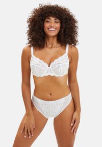 Soutien-gorge en dentelle blanche avec des bords festonnés et des motifs floraux, associé à une culotte semi-transparente assortie présentant des détails en dentelle similaires.