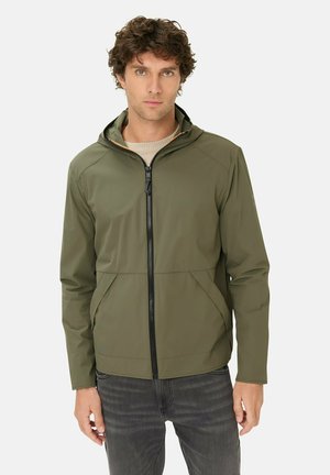 Man met krullend haar, gekleed in een olijfgroene zip-up capuchontrui en donkergrijze spijkerbroek, staand tegen een effen witte achtergrond.