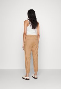 Beige elastic-waist trousers with tapered legs, two back pockets, and a smooth fabric. Paired with a white tank top and light sandals.