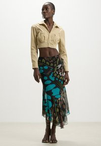 Maglietta beige cropped con dettagli arricciati, abbinata a una lunga gonna trasparente multicolore con motivi di farfalle blu e nere.