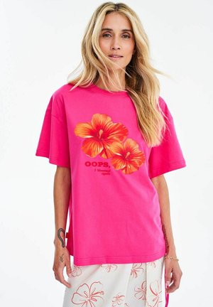 Blondynka ubrana w jasnoróżowy, oversize T-shirt z dwoma dużymi kwiatami hibiskusa i napisem, w zestawieniu z białymi spodniami w kwiatowy wzór.