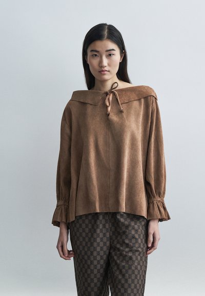 Blouse en daim marron avec un col large et replié et un détail noué au niveau du cou. Manches longues avec des poignets à volants. Portée avec un pantalon marron à motifs.