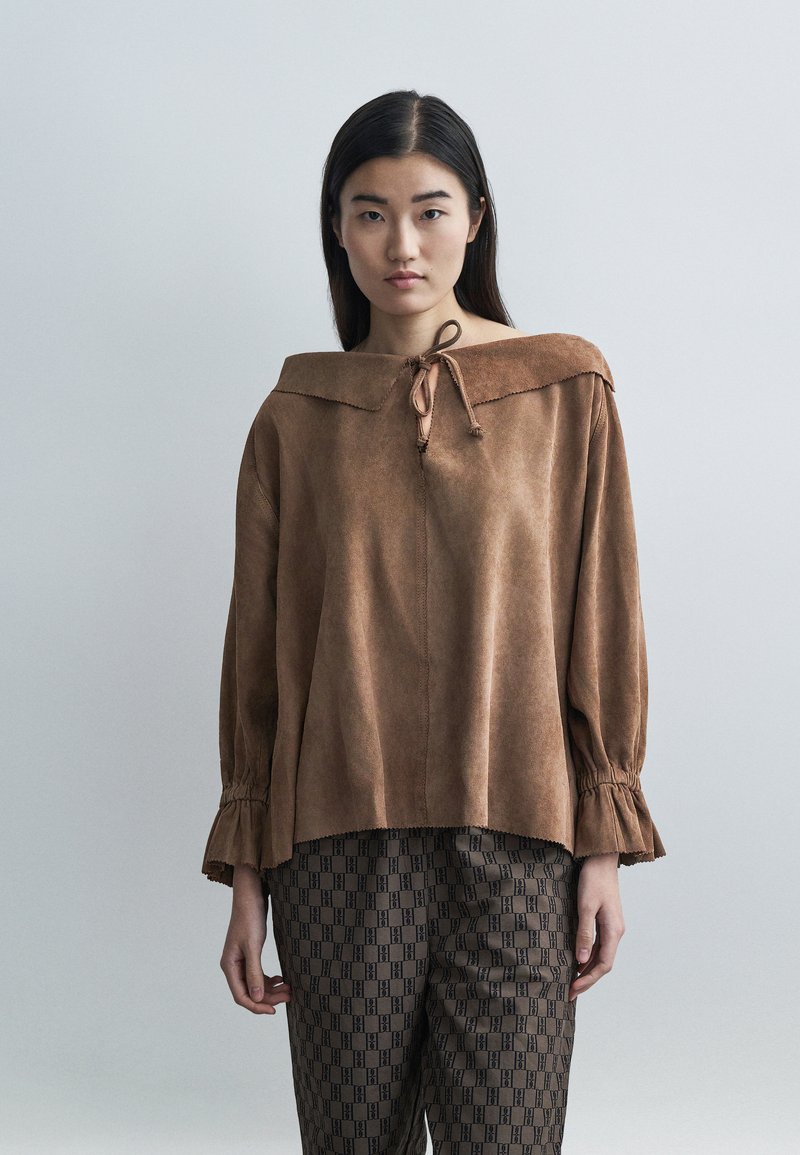 Καφέ μπλούζα από σουέντ με široκλο και διπλωμένο γ�ιακά και δεμένο λαιμόκοψη. Διαθέτει μακριά μανίκια με μανσέτες με βολάν. Φορεμένη με καφέ παντελόνι με μοτίβο.