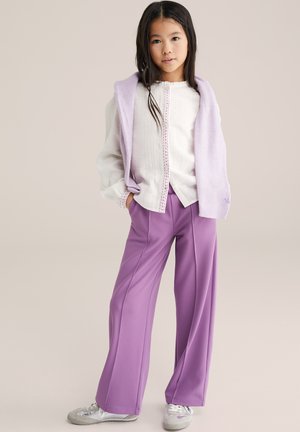 Niña joven de pie vistiendo blusa blanca, suéter lila sobre los hombros, pantalones anchos morados y zapatillas blancas.