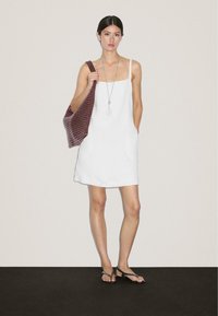 Frau in einem weißen ärmellosen Kleid, die eine große kastanienbraune strukturierte Tasche hält, eine lange silberne Halskette und schwarze Sandalen trägt und auf einem schwarzen Boden steht.