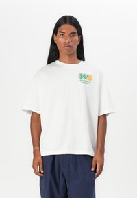 Fehér, oversized póló, kerek nyakkivágással, zöld és narancssárga grafikával, amelyen a "Worldwide Hoops" felirat látható a mellrészen.