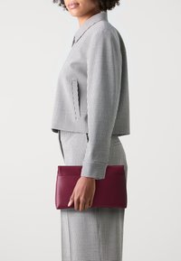 Porte-monnaie en cuir bordeaux tenu par une personne, associé à un blouson court à carreaux noir et blanc et à un pantalon assorti. Texture lisse.