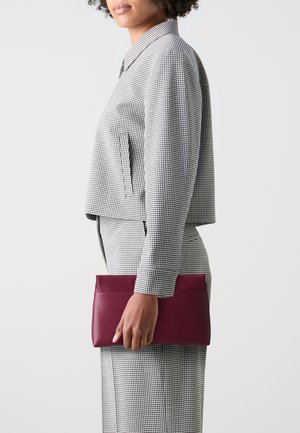 Porte-monnaie en cuir bordeaux tenu par une personne, associé à un blouson court à carreaux noir et blanc et à un pantalon assorti. Texture lisse.