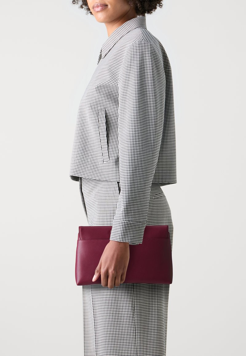 Porte-monnaie en cuir bordeaux tenu par une personne, associé à un blouson court à carreaux noir et blanc et à un pantalon assorti. Texture lisse.