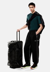 Borsa da palestra nera con ruote, dal texture liscia, con fibbie e logo EA7. Il modello indossa pantaloni scuri, una maglietta verde e scarpe nere con dettagli verdi.