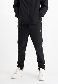 Zwarte sportbroek gemaakt van lichtgewicht materiaal, met een relaxte pasvorm, zijzakken en subtiele branding op de dij.