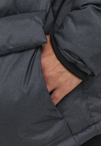Mørkegrå polstret jakke med struktureret finish, der har en hånd i en sidelomme og en sort elastisk manchet-detail.