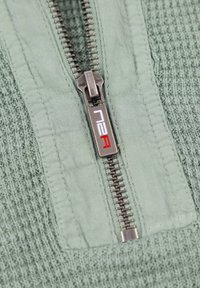 Lichtgroene gemêleerde stof met een reeks horizontale ribbels, met een zilveren ritsdetail en een logo met rode accenten op de pulltab.