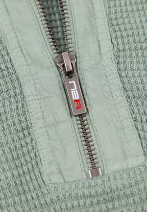 Lichtgroene gemêleerde stof met een reeks horizontale ribbels, met een zilveren ritsdetail en een logo met rode accenten op de pulltab.