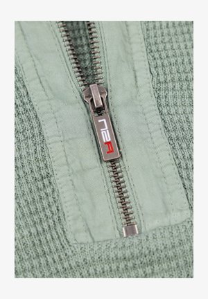 Lichtgroene gemêleerde stof met een reeks horizontale ribbels, met een zilveren ritsdetail en een logo met rode accenten op de pulltab.