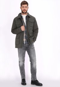 Olijfgroene jas met meerdere voorzakken, knoopsluitingen en een kraag; gedragen over een geruit overhemd en grijze spijkerbroek, gecombineerd met zwarte laarzen.