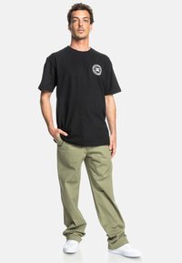 Sort bomulds t-shirt med et cirkulært logo på brystet, kombineret med olivengrønne bukser og hvide sneakers. Afslappet pasform, glat tekstur.