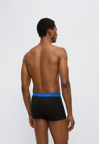 Μαύρα ανδρικά εσώρουχα boxer με μπλε ζώνη μέσης που φέρει το λογότυπο «Tommy Hilfiger», απεικονιζόμενα από πίσω σε ουδέτερο φόντο.