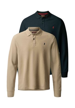 Homme avec barbe et cheveux ondulés porte un polo bleu marine à manches longues, un pantalon bleu foncé et des bottes en cuir marron, debout avec une main dans la poche.