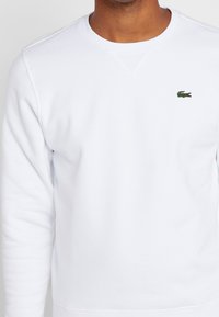 Vit sweatshirt med rundad halsringning och långa ärmar, prydd med en liten grön krokodil-logotyp på vänster bröst. Mjuk tygtextur.