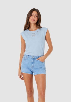 Donna che indossa un top azzurro senza maniche con scritta e pantaloncini di jeans azzurri, in piedi con una mano in tasca su uno sfondo semplice.