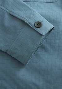 Chemise bleu clair avec un motif texturé en forme de losanges, poignets boutonnés et finition en tissu lisse.