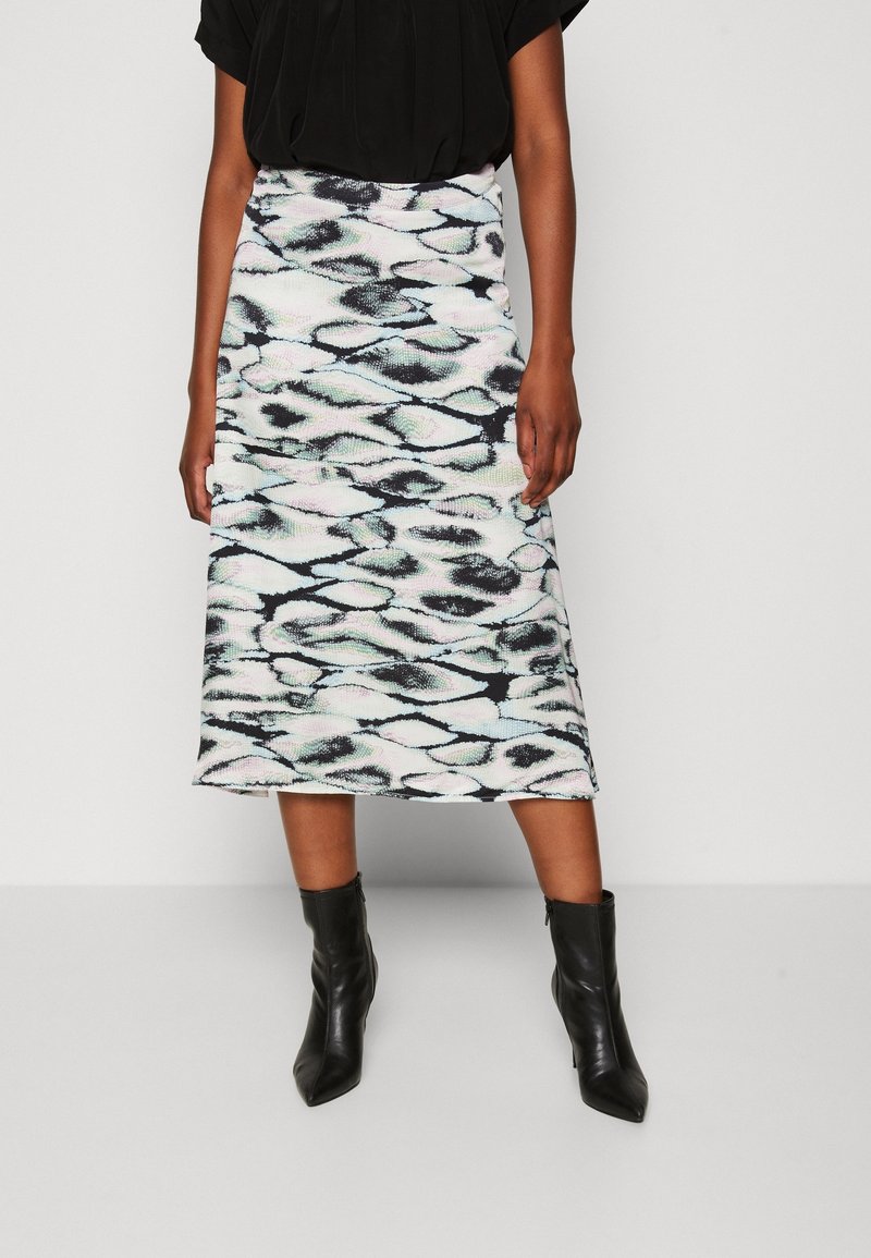 Femme portant un haut noir, une jupe mi-longue avec un motif abstrait noir et blanc, et des bottines noires, debout devant un fond uni.