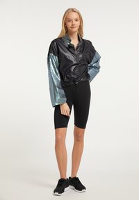 Sort zip-up jakke med blanke sølv-blå mønstrede ærmer, parret med sorte cykelshorts og sorte atletiske sko på en lys baggrund.