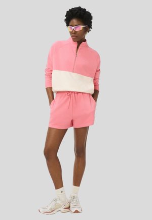 Vrouw draagt een roze en crème sweatvest met rits, bijpassende roze shorts, witte sneakers, witte sokken en reflecterende sportzonnebril, staand met handen in de zakken.