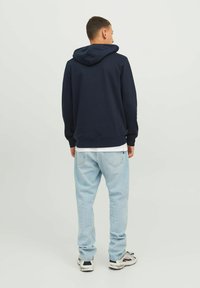 Sudadera azul marino con capucha ajustable, de corte relajado, llevada sobre una camiseta blanca, combinada con vaqueros de un tono azul claro y zapatillas negras.