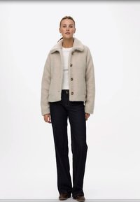 Beige fleecejacka med krage, tre knappar och en avslappnad passform, tillsammans med mörka denimjeans och bruna skor. Slät textur.