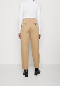 Pantaloni beige con vestibilità sartoriale, dotati di tasche posteriori, una texture liscia e gamba leggermente svasata. Abbinati a un top bianco a maniche lunghe.