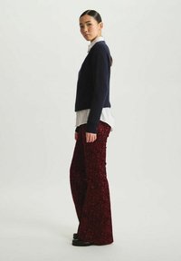 Pull marinier superposé à une chemise blanche ; pantalon à pattes d'eph rouges avec un motif floral texturé ; modèle portant des chaussures noires, se tenant de côté.