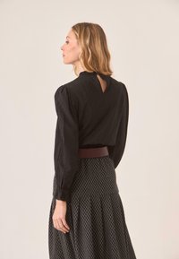 Blusa negra de manga larga con cuello con volantes y detalle de ojal en la parte posterior, combinada con una falda negra estampada y un cinturón burdeos.