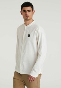 Camicia bianca a maniche lunghe con bottoni, realizzata in un tessuto testurizzato, dotata di colletto alla cinese e di una piccola toppa nera con logo sul petto.