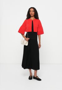 Giacca corta rossa con maniche a campana, abbinata a una gonna nera a pieghe. La modella porta anche una piccola borsa color crema e indossa scarpe nere intrecciate.