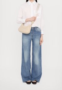 Borsa a tracolla in pelle beige con forma arrotondata, dotata di una tracolla robusta e un marchio discreto. Abbinata a una camicia bianca con bottoni e jeans a gamba larga.